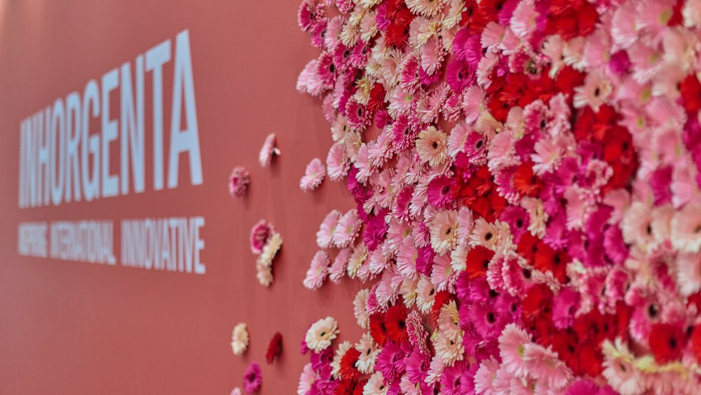 Wand mit „INHORGENTA“-Schriftzug und bunten Blumen, vorwiegend in Rosa und Rot, in einem Blumenarrangement arrangiert.