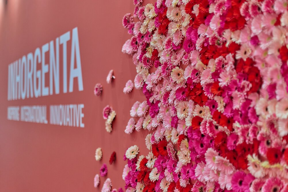 Wand mit „INHORGENTA“-Schriftzug und bunten Blumen, vorwiegend in Rosa und Rot, in einem Blumenarrangement arrangiert.