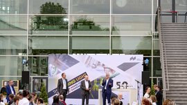 Eine Gruppe von Menschen vor einer Bühne vor einer Messehalle an der drei Redner gerade eine Preisverleihung ankündigen.