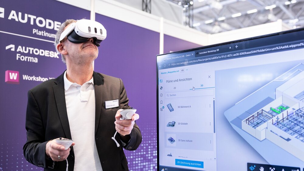 Ein Mann trägt eine VR-Brille und hält zwei Steuerelemente in den Händen. Im Vordergrund ist ein Bildschirm zu sehen, der eine Baustelle zeigt.