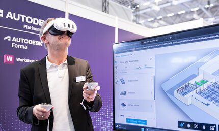 Ein Mann trägt eine VR-Brille und hält zwei Steuerelemente in den Händen. Im Vordergrund ist ein Bildschirm zu sehen, der eine Baustelle zeigt.