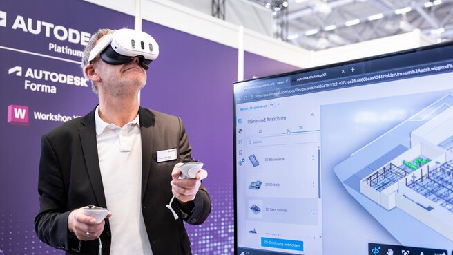 Ein Mann trägt eine VR-Brille und hält zwei Steuerelemente in den Händen. Im Vordergrund ist ein Bildschirm zu sehen, der eine Baustelle zeigt.