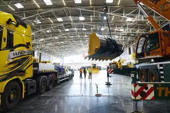 A large yellow excavator bucket is delivered to an exhibition hall by crane. Several workers in protective clothing watch the assembly.