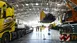 A large yellow excavator bucket is delivered to an exhibition hall by crane. Several workers in protective clothing watch the assembly.