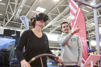 Eine Frau probiert an einem Messestand eine VR-Brille aus und hält ein Steuerrad in der Hand. Rechts steht ein Mann, der ihr das Erlebnis erklärt.