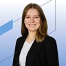 Eine Frau in einem schwarzen Blazer und einem weißen Hemd, die vor einem blauen geometrischen Hintergrund lächelt.