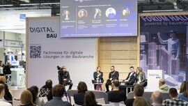 Fünf Personen sitzen auf einer Bühne unter einer großen Leinwand, auf der ihre Namen und die Messe: digitalBAU 2024, stehen. Mehrere Personen hören dem Vortrag zu.