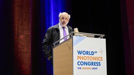 An older man with a purple tie and red glasses speaks on the podium of the World of Photonics Congress.