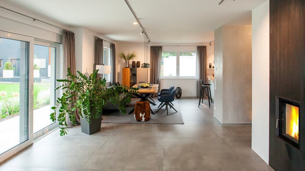 Moderner offener Wohnbereich mit großen Fenstern, Esstisch und Grünpflanzen. Kamin an der rechten Wand.