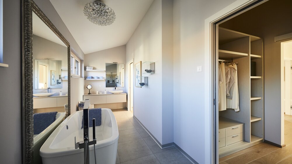 Modernes Badezimmer mit freistehender Badewanne, großem Spiegel und offenem begehbarem Kleiderschrank. Neutrale Töne und Tageslicht.