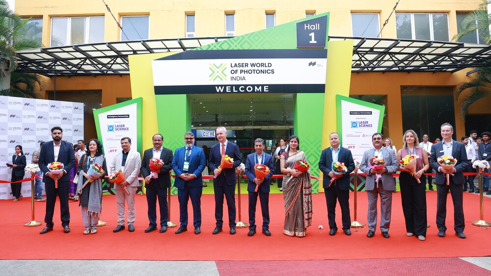 Several people in festive attire and holding bouquets of flowers pose for a photo in front of the entrance to Laser World of Photonics India.