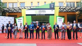 Several people in festive attire and holding bouquets of flowers pose for a photo in front of the entrance to Laser World of Photonics India.
