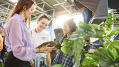 Drei junge Frauen unterhalten sich lächelnd auf einer Messe, umgeben von Pflanzen und Dachzelten in heller, moderner Halle.