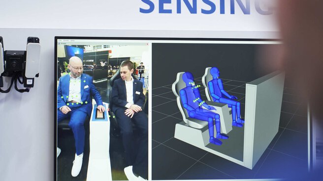 A display on the wall shows software that records the sitting position of two people with sensors and displays it as a 3D model.