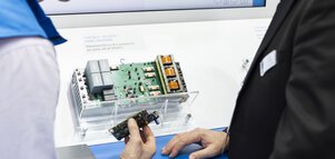 Two men stand in front of an energy conversion module on display at a trade fair stand. The man on the left is holding a small circuit board.