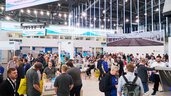 Blick in eine belebte Messehalle der Firma Huber mit zahlreichen Besuchern und herabhängenden LED-Lichtstäben an der Decke.