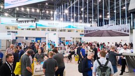 View into a busy exhibition hall of the Huber company with numerous visitors and LED light tubes hanging from the ceiling.