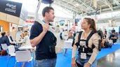 Two people in technical equipment are talking in a busy exhibition hall with stands and seated participants in the background.