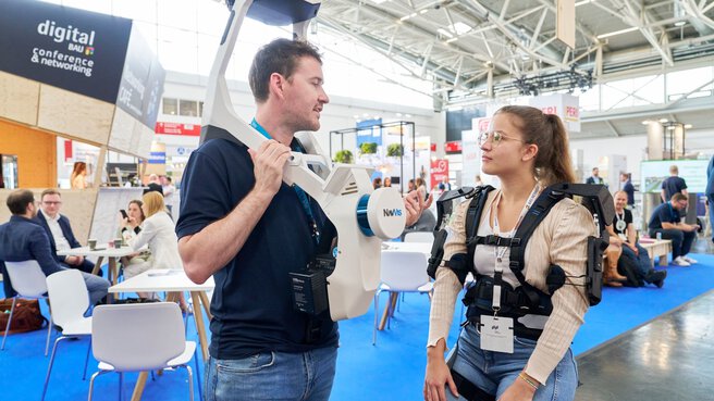 Two people in technical equipment are talking in a busy exhibition hall with stands and seated participants in the background.
