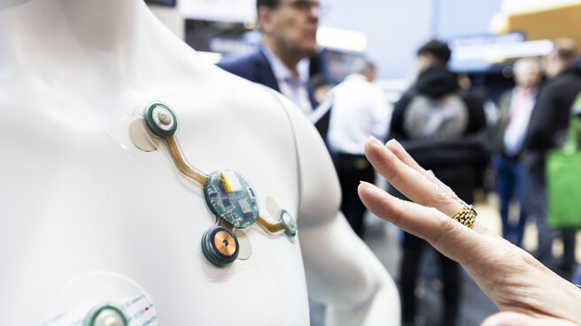 A hand stretched out in front of the chest of a mannequin with electronic circuits and sensors glued to it. Visitors and exhibition stands in the background.