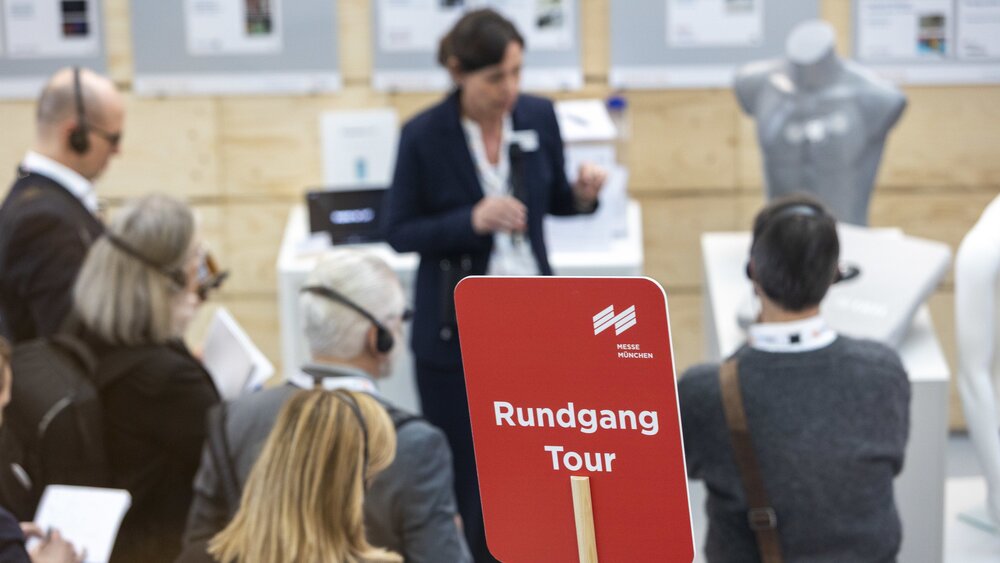 Ein großes rotes Schild mit dem Text „Rundgang Tour“ vor einer Gruppe Messebesucher, die über Kopfhörer den Ausführungen einer Frau zuhören.
