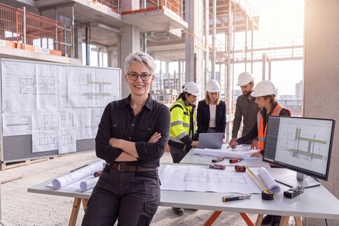 A woman is leaning against a desk on a building site; in the background, several people are standing in workwear