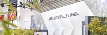 People gathered around tables at an exhibition stand. In the background is a coffee bar and a large sign with the words “Efficiency in all ways”.