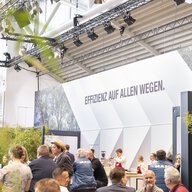 Menschen versammelten sich um Tische an einem Messestand. Im Hintergrund ist eine Kaffeebar und ein großes Schild mit der Aufschrift „Effizienz auf allen Wegen“.