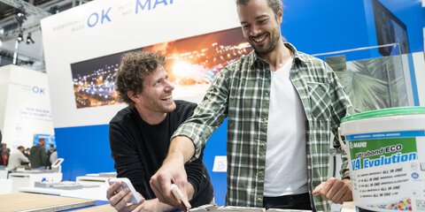 Zwei lächelnde Männer stehen gemeinsam an einem Stand auf der BAU und testen mit einem Spachtel einen Universalklebstoff.