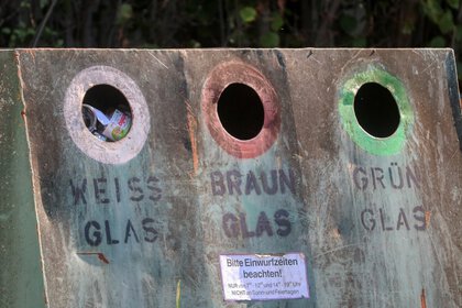 Nahaufnahme eines Altglascontainers mit drei runden Einwurföffnungen für Weiß-, Braun- und Grünglas; unter den Öffnungen sind die jeweiligen Beschriftungen sowie ein Hinweis zu den Einwurfzeiten zu sehen.