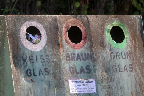 Close-up of a glass recycling container with three round openings for white, brown, and green glass; the respective labels and information on recycling times can be seen below the openings.