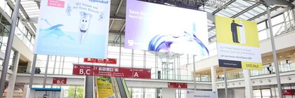 Eingangshalle eines Messezentrums mit dicht gedrängten Besucherinnen und Besuchern vor Rolltreppen, über denen große Werbe- und Hinweistafeln für Aussteller und Messebereiche hängen.