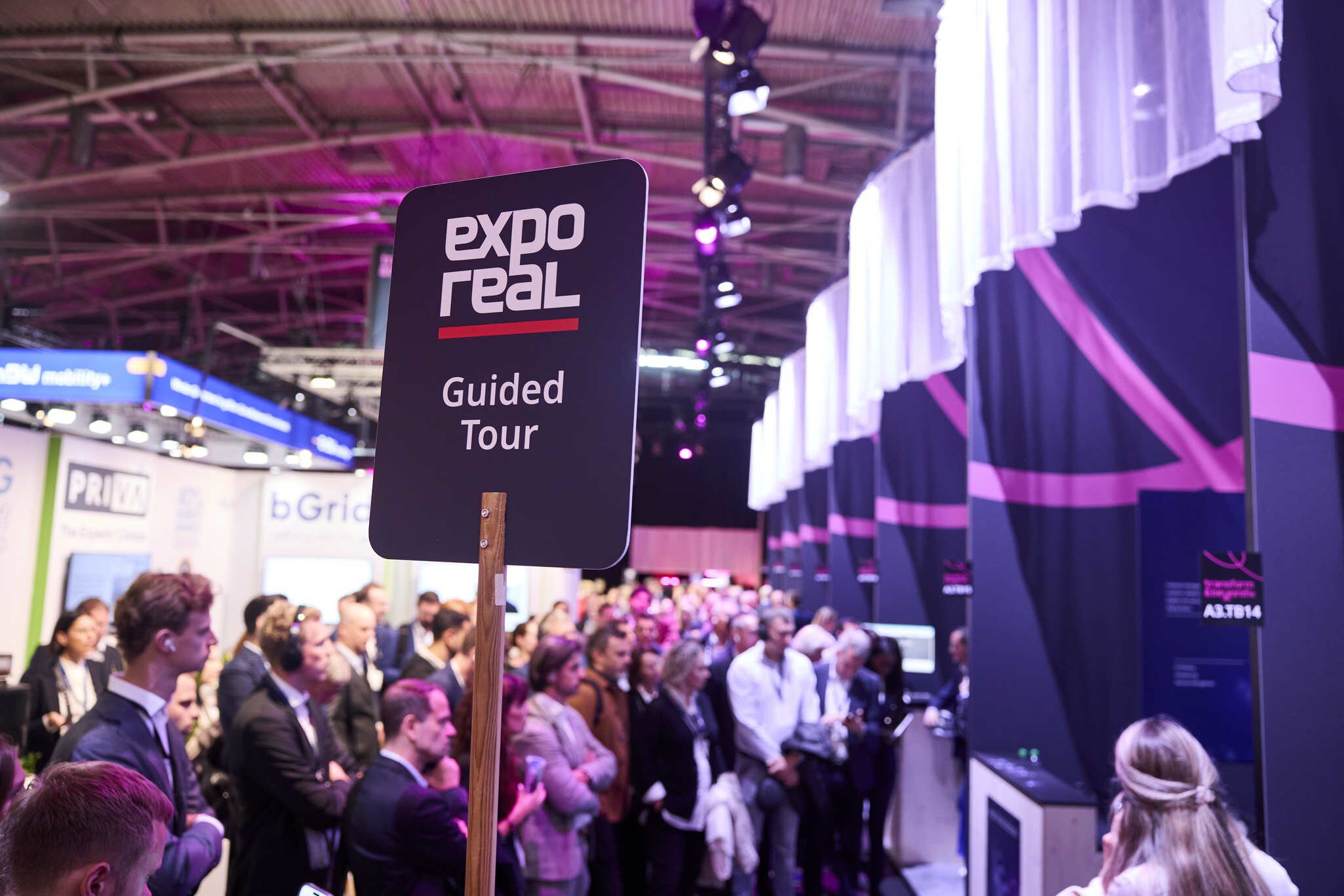 A group of people with headphones gather in front of an exhibition stand. A sign reading “Guided Tour” is held up in the foreground.