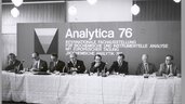 Schwarz-weiß Foto. Männer sitzen hinter einem langen Tisch mit Miktofonen und geben eine Pressekonferenz. Im Hintegrund steht auf einer Stellwand: Analytica 76 - Internationale Fachausstellung für biochemische und instrumentelle Analyse mit europäischer Tagung.