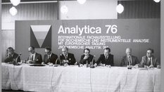 Black and white photo. Men are sitting behind a long table with miktophones and giving a press conference. In the background, a billboard reads: Analytica 76 - International Exhibition for Biochemical and Instrumental Analysis with European Conference.