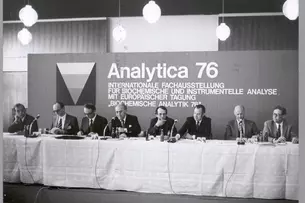 Black and white photo. Men are sitting behind a long table with miktophones and giving a press conference. In the background, a billboard reads: Analytica 76 - International Exhibition for Biochemical and Instrumental Analysis with European Conference.