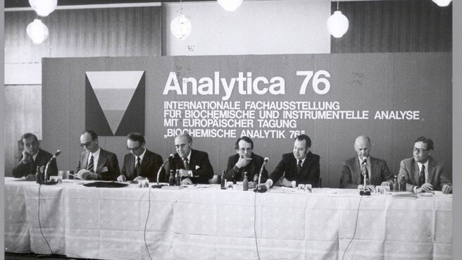 Schwarz-weiß Foto. Männer sitzen hinter einem langen Tisch mit Miktofonen und geben eine Pressekonferenz. Im Hintegrund steht auf einer Stellwand: Analytica 76 - Internationale Fachausstellung für biochemische und instrumentelle Analyse mit europäischer Tagung.