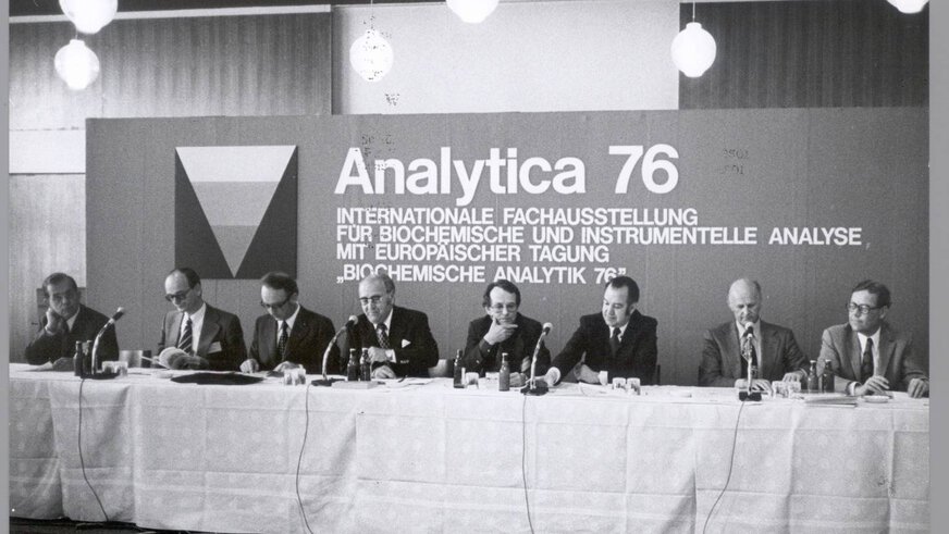 Black and white photo. Men are sitting behind a long table with miktophones and giving a press conference. In the background, a billboard reads: Analytica 76 - International Exhibition for Biochemical and Instrumental Analysis with European Conference.