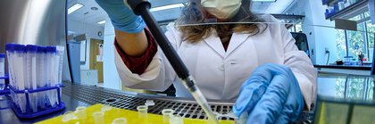 A person in a lab coat and medical mask uses a pipette on a yellow dish with samples in tubes.