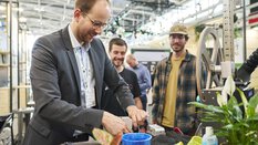 Ein Mann demonstriert an einem Messestand vor zwei Besuchern die Handhabung eines Geräts.
