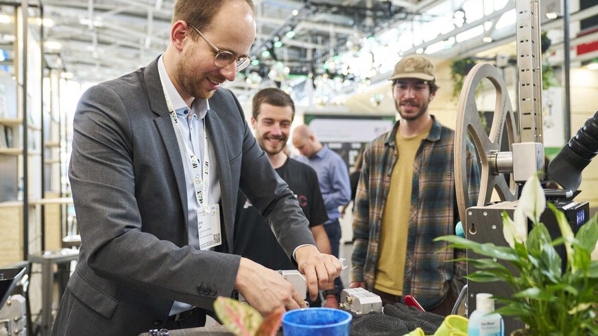 Ein Mann demonstriert an einem Messestand vor zwei Besuchern die Handhabung eines Geräts.