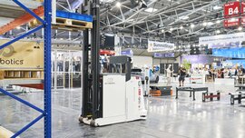 Autonomous transport vehicle in an exhibition hall with shelving and other robots.