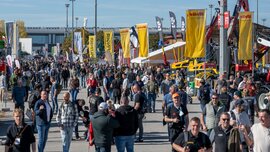 Eine große Menschenmenge, die durch eine Industrieausstellung im Freien mit vielen Fahnen, Transparenten und schweren Baumaschinen schlendert.