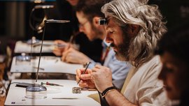 Several people are seated in a row at tables, working on watch cases with delicate tools. The man in focus is wearing a watchmaker's loupe on his forehead.