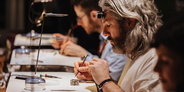 Several people are seated in a row at tables, working on watch cases with delicate tools. The man in focus is wearing a watchmaker's loupe on his forehead.