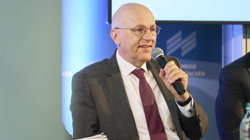 A man in a suit speaks into a microphone at an event. The Messe München logo can be seen blurred in the background.
