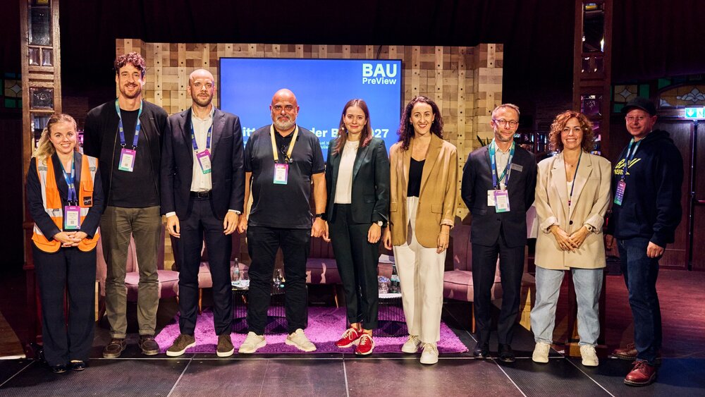 Eine Gruppe von zehn Personen mit Konferenzbadges steht auf einer Bühne vor einem BAU-PreView-Bildschirm und blickt für ein gemeinsames Foto in die Kamera.