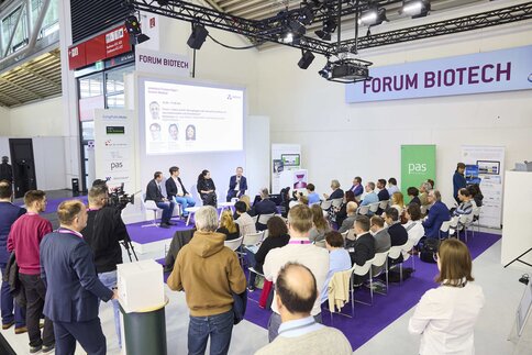 The audience is watching a panel discussion on biotechnology on the conference stage, whilst the speakers sit beneath a large screen.