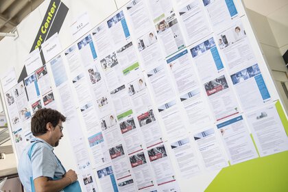 Stellwand die mit ausgedruckten Fachartikeln im A4 Format behängt ist. Ein Besucher steht davor und liest einen Artikel.