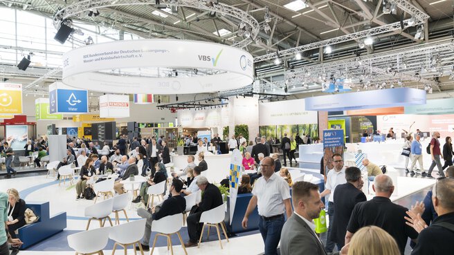 Visitors to a trade fair on the subject of waste disposal, water and recycling management. The focus is on discussions and networking in an inviting lounge atmosphere. The logos of exhibitors such as BDE and VBS can be seen above the stands. In the background, various companies and associations present sustainable solutions and innovations.